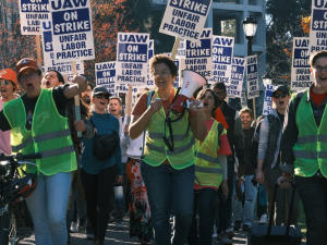 Strikers march outdoors. The three in front, wearing neon vests of picket captains, are chanting energetically, two with fists in air, one holding bullhorn. Crowd behind them holds "UAW on strike" picket signs.