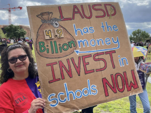 A woman in a red UTLA t-shirt holds a cardboard sign with red, blue, white and green lettering showing a big bag of money and saying “LAUSD has the money, 4.9 billion, invest in schools now.” She’s standing in a field with other demonstrators in the background. 