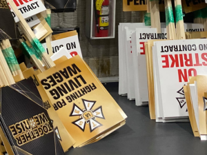 Picket signs stacked against a wall. They say "FIGHTING FOR LIVING WAGES," "ON STRIKE NO CONTRACT," and "TOGETHER WE RISE" and have the IATSE logo. There is also a fire extinguisher in the photo.