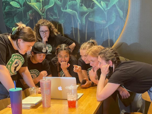 Young workers lean over a table, all peering intently at one laptop screen. They look excited and happy.