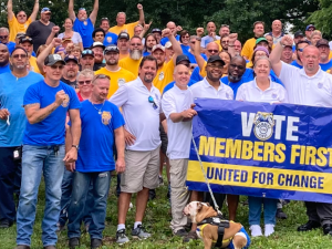 Big crowd stands on grass outdoors on a sunny day behind a banner that reads "Vote Members First, United for Change." Everyone is wearing a union shirt: white, blue, or yellow. Many have fists in the air and everyone is smiling. Someone in front has a dog on a leash.