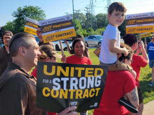 Workers picket with "United for a strong contract" and "Just practicing" teamsters signs. Some wear brown UPS driver uniforms. One little kid, riding on a parent's shoulders, turns to look happily directly into the camera.