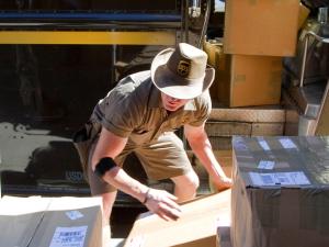 A uniformed UPS delivery driver, wearing a UPS-branded cowboy hat, lifts large packages onto a rolling cart in front of his delivery truck. More large packages are visible through the truck's open door.