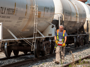 A worker in reflective vest walks alongside a train, holding a remote control device