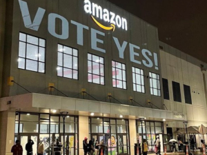 Warehouse lit up against dark sky. Directly under the "Amazon" logo the message "Vote yes!" is projected on the wall. A few people are visible coming and going near the doors below.