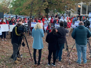 A crowd of red-shirted people stands outside Lawrence High School. One person is speaking to the group; others are shooting video of the event.