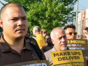 Crowd in UPS brown uniforms stands outdoors looking resolute, some holding signs. One sign visible reads "Make UPS Deliver!"