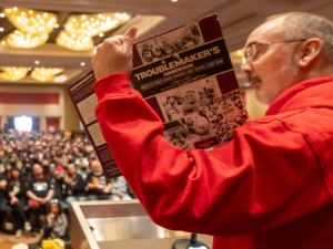 Seen from the side/back, Shawn Fain in a red hoodie holds up a battered purple copy of A Troublemaker's Handbook. Arrayed in front of him can be seen a huge seated crowd in the ballroom for the closing plenary.