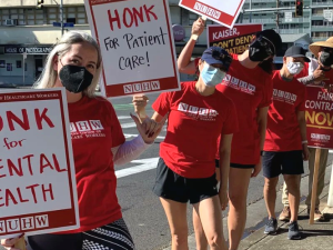 Six picketers in red NUHW T-shirts stand on outdoors along a sidewalk. Most are masked. Some carry handmade signs that say "Honk for mental health" and "honk for patient care." Others carry printed signs that say "Fair contract now" and "Patient health not corporate wealth."