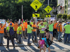 Big crowd of carpenters in high-vis neon orange and green shirts stands on a street corner. Some hold handmade signs reading "PAY UP AGC" and "VOTE NO."