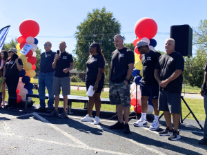 About a dozen adults and a couple children, white and Black, men and women, stand mostly silhouetted in bright sun. Most wear black union T-shirts.They're outdoors, on pavement in front of grass. One holds a mic. Some hold printed signs that say, "IUE-CWA, Better Wages." There are also two columns of balloons, each a spiral design in white, red, yellow, and black.