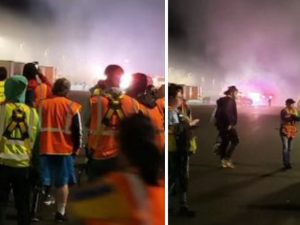 People in neon yellow and orange vests stand in front of clouds of purple smoke outside a warehouse at night.