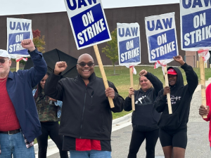 Six smiling workers carrying "UAW On Strike" picket signs raise their fists in the air outside a GM CCA facility. The workers are a mix of Black and white, men and women. Some of the picket signs are wrapped in plastic bags, and one worker carries an open umbrella.