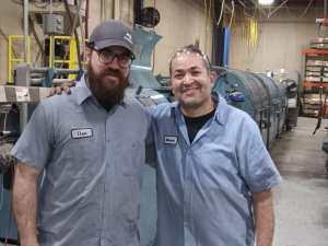 Two men in blue work shirts with nametags "Dan" and "Mohamed" sewn on stand side by side, arms around each other's shoulders, in a machine shop