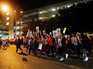 Workers with "On Strike" picket signs march past a hospital in the near-dark, silhouetted by one bright light.