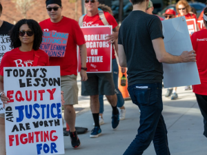 Strikers picket in red shirts. Most prominent is a woman with a sign: "Today's lesson: equity and justice are worth fighting for." Behind her is a man with a partially obscured sign listing "Part-time jobs I've worked so I can afford to be a teacher in Massachusetts" including Lyft driver, nightclub bouncer, Doordash, adjunct. 