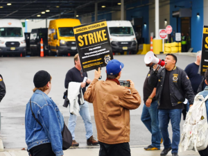 Workers picket in front of an Amazon facility where delivery vehicles are visible. Some carry printed "Amazon workers on strike" signs with a Teamsters logo. Others wear satin jackets with a Teamsters logo, and one shouts into a bullhorn.