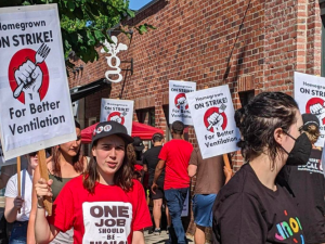 People picket in a circle outside a brick building. Their printed signs say "Homegrown on Strike! For Better Ventilation" with a logo of a fist holding a fork. Person in the foreground wears a red UNITE HERE shirt with the slogan "One job should be enough."