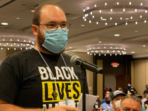 white man in mask at mic in hotel banquet room, wearing "Black Lives Matter" shirt, speaks as other people look on