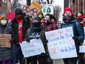 Crowd in coats and masks. One hand-drawn sign reads "Roses are red, educators are blue, if one of us dies, Kenney, what will you do?" Another shows a train and reads "Choo-choo-choose a safe plan." Another says "ventilate, vaccinate, educate." Another shows an image of a fan with a "no" line across it.