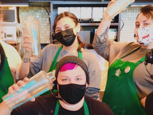 Six young workers in a Starbucks, wearing the trademark green aprons, look cheerfully or defiantly at the camera. Three are holding cups aloft.