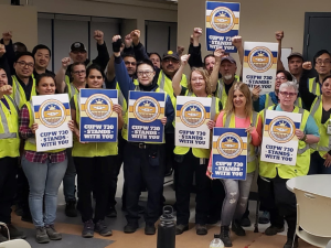 A group of smiling people in yellow vests stands inside a cafeteria. Several hold signs "CUPW Local 730 stands with you." Many have their fists in the air.