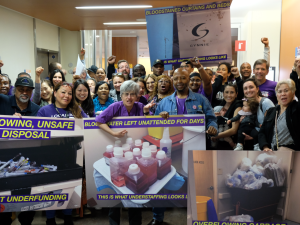 Big crowd of members in a hallway, many fists in air. Front row holds oversized photo signs with captions: "OVERFLOWING, UNSAFE SHARPS DISPOSAL: THIS IS WHAT UNDERFUNDING LOOKS LIKE." "BLO{***}ATER LEFT UNATTENDED FOR DAYS: THIS IS WHAT UNDERSTAFFING LOOKS LIKE." "PATIENTS DISCHARGED IN FILTHY CLOTHING."]