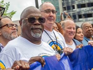 Black and white people, including UAW President Shawn Fain, hold a blue banner (text not visible). They are wearing matching T-shirts for a Martin Luther King Official Commemoration.