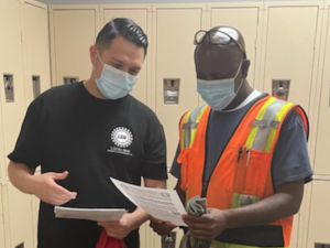 Two men talk inside an auto plant locker room, looking together at leaflets. Both wear masks and one wears an orange reflective vest.