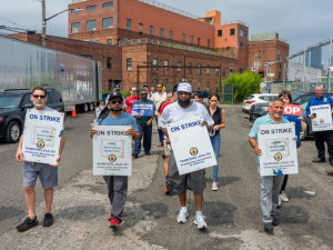 Workers walk forward outdoors carring signs reading "ON STRIKE: United Metro Energy Company: Teamsters Local 553." One person has a hand-lettered sign: "Teacher solidarity" that is flopping in the wind. Behind them is a brick industrial building. 