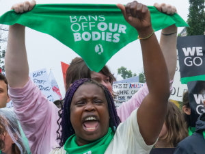 Women shout and raise their fists at a rally. Many are wearing green bandannas and one is holding the bandanna up in the air: it says "Bans off our bodies." A sign held up in background says "We won't go back."