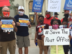 Masked people (Black and white, men and women) holding signs. Most prominent is a handwritten sign: "#Save the Post Office." Preprinted signs say "Stand with Postal Workers" and "Senators, do the right thing! Save USPS"