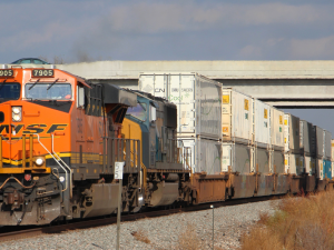 A freight train passes along a track under an overpass. The orange engine is labeled BNSF. It is pulling many white freight cars.