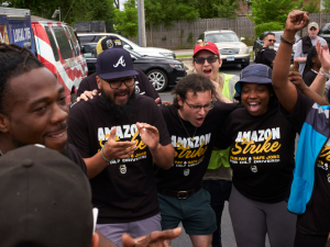 A young, racially diverse group of workers in "Amazon Strike" T-shirts huddles in a parking lot together, cheering. Some raise fists in the air. Behind them a Teamsters Local 705 truck is visible.