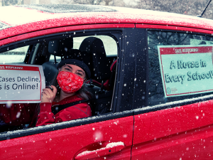 Person in driver's seat of red car, wearing red mask, holds sign: "Safe reopening! Until cases decline, class is online!" Taped to back window of car is another sign: "A nurse in every school!" It is snowing.