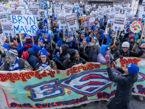 Huge, shouting crowd of people in blue hats, many with picket signs "On strike for smaller class sizes." One prominent handmade sign says "Bryn Mawr." A big banner at front of crowd says, in artsy spray-painted style, "District refuses to pay for ESPs, our children's education heroes!" The S in ESPs is the Superman logo. 