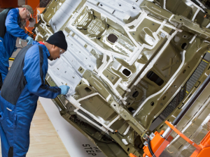 Two workers in blue overalls lean over a car body, doing detailed work