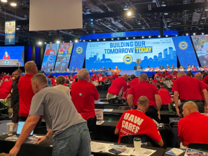 People in a big convention hall, all wearing red T-shirts, most getting to their feet. They are viewed from the back. At the front of hte room is a dais with people seated, and a huge lighted display that says "Building our tomorrow today" with the UAW logo.