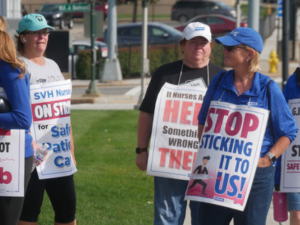 Several nurses picket outside wearing sign. One sign says "Stop sticking it to us!" with a drawing of a person in a business suit puncturing the letters with a giant syringe and needle. Another says "G.I. am tired of [poop emoji] staffing. SAFE STAFFING NOW. -Endoscopy." Other signs are partially obscured but appear to say "Don't lie, we can spot A-Fib," "If nurses are out here there's something wrong in there," and "SVH nurses on strike for safe patient care."