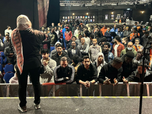 A large crowd of workers in a dark auditorium listen to a worker onstage. The speaker's back is to us and she wears a head scarf or hijab. Some in the audience are smiling, some look focused, and two in the front row are animatedly discussing something. The workers are racially diverse, including many who could be of South Asian or East African descent, as the article describes.