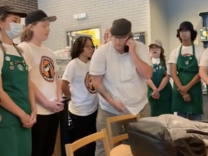 Young workers in Starbucks aprons and SBWU logo shirts stand in a semicircle, all in calm postures mostly with hands clasped at rest in front of them. In the center is the manager in a white shirt, talking into a cell phone.