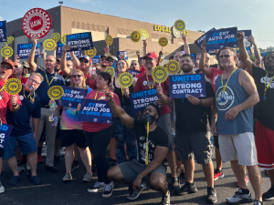 An energetic crowd of workers of various ages, genders, and races, Shawn Fain among them, poses in the sunshine outside a building marked Local 862. Many hold printed signs: "United for a strong contract," "Every job a good job," and the UAW logo. Many thrust their fists in the air.