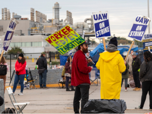 Workers in heavy coats stand around picketing in front of a big gray factory. One neon-colored handmade sign says "Worker-led EV transition" with a lightning bolt. Other signs are printed and say "UAW on strike" or "UAW, Stand up, Record Profits, Record Contracts" 