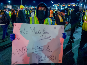 Person in yellow vest at night, outdoors in crowd, holds handwritten sign: "We make you millions, we want a dollar!"