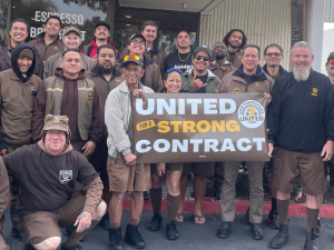 26 people (mostly men) in brown UPS uniforms pose outside Duke's coffee shop, smiling. Some hold a banner: "United for a strong contract" with the UPS Teamsters United logo.