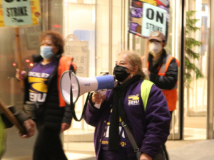 People in masks and purple SEIU 87 shirts picket in front of a bright window. Central person has a bullhorn. Others carry "ON STRIKE" signs and wear reflective vests.