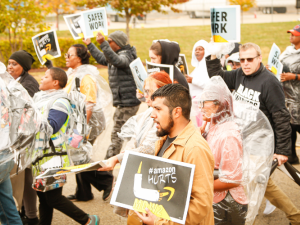 Workers, many in ponchos, march briskly on a street. Some carry printed signs saying "Safer work," "Higher wages," or "Amazon hurts." 