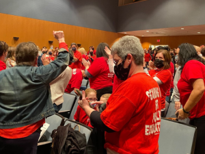 People, mainly women, in red T-shirts in the process of getting up en masse to exit an auditorium. One has a fist in the air, and several are clapping. Most are viewed from the back or side. The backs of their T-shirts say "STANDING UP FOR PUBLIC EDUCATION"