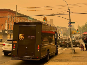 UPS truck viewed from behind on a Brooklyn street; visible are a few pedestrians and a fruit market. The sky and the whole scene is blanketed in a thick orange haze; buildings down the block are fuzzier because of the smoke.