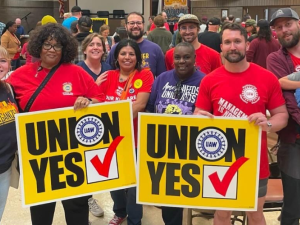 Ten smiling people pose together in a crowded meeting room. Two are holding huge yellow "Union Yes" signs with the UAW logo. Most are wearing red, blue, or purple T-shirts with UAW logo or organizing-themed slogans. The man on the far right holds a small child in his arms.